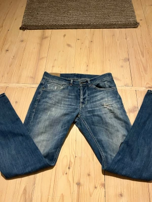 Blå jeans från dondup med slitningar och detaljer - Snygga blå jeans från dondup med slitna detaljer och ljusa partier för en cool, använd look. Klassisk femficksmodell med knapp. Jeansen har kontrastsömmar och en liten broderad detalj på bakfickan. Perfekta för en avslappnad streetstil.