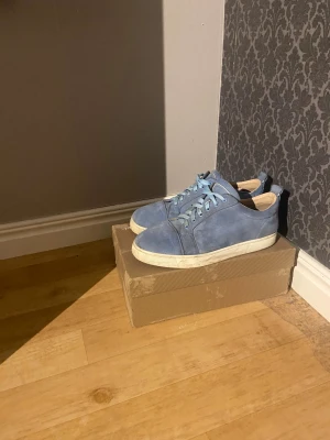 Louboutin - Snygga blå sneakers i mocka från Christian Louboutin med klassisk röd sula och rund tå. Skorna har snörning och vit platt sula, vilket ger en clean och modern vibe. Perfekt för dig som vill sticka ut med exklusiva detaljer och lyxig känsla.