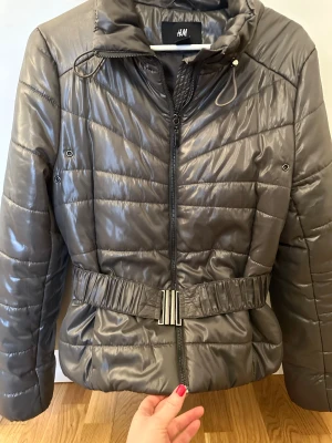 Grå pufferjacka från H&M med bälte - Snygg grå pufferjacka från H&M i storlek 36/38. Jackan har quiltad design, hög krage, dragkedja framtill och ett brett bälte i midjan med metallspänne. Materialet är glansigt.. Perfekt för  🍂 hösten.