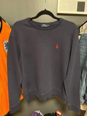 Mörkblå sweatshirt från Polo Ralph Lauren - Snygg mörkblå sweatshirt från Polo Ralph Lauren med klassisk röd logobrodyr på bröstet. Tröjan har rund hals, ribbade muddar och långärmad passform. Perfekt för dig som gillar stilrena och tidlösa plagg med en sportig touch. Storleken är M i barnstorlek!