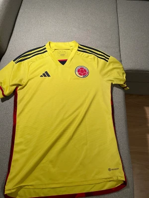 Colombia landslagströja Adidas gul M - Gul Colombia fotbollströja från Adidas med broderat landslagsemblem och mörkblå Adidas-logga. Tröjan har svarta ränder på axlarna, röda detaljer på sidorna och är tillverkad i ventilerande Aeroready-material. Perfekt för dig som älskar fotboll och vill sticka ut.