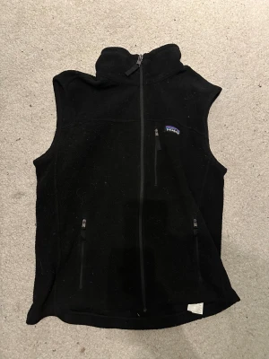 Patagonia fleeceväst - Svart fleeceväst från Patagonia med hög krage och hel dragkedja framtill. Västen har tre fickor med dragkedja och en klassisk logga på bröstet. Perfekt för lager-på-lager och outdoor vibes.