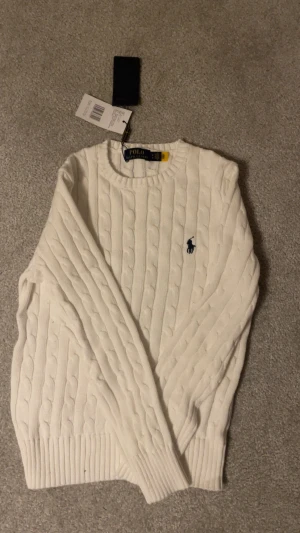 Vit kabelstickad tröja Polo Ralph Lauren - Snygg vit kabelstickad tröja från Polo Ralph Lauren med klassisk rund hals och marinblå broderad logga på bröstet. Tröjan har långa ärmar och ribbade muddar vid ärmslut och nederkant. Perfekt för dig som gillar stilrena och tidlösa plagg.