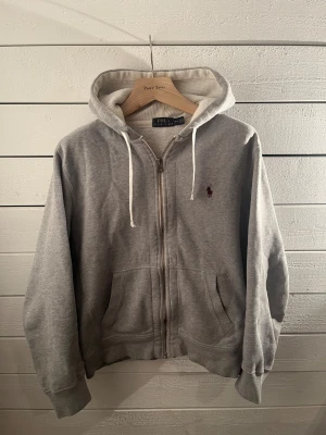 Grå hoodie från Polo Ralph Lauren - Snygg grå hoodie från Polo Ralph Lauren med dragkedja, huva och vita snören. Tröjan har ett broderat vinrött Polo-logo på bröstet och två praktiska fickor framtill. Mjuk insida och klassisk passform, perfekt för chill dagar.