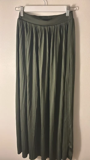 Olivgrön plisserad långkjol About You - Snygg olivgrön plisserad långkjol från About You. Kjolen har ett brett midjeband och faller i mjuka veck för en flowig look. Perfekt att styla med sneakers eller boots för en trendig vibe.