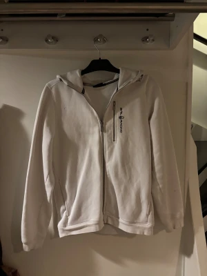 Vit hoodie från Sail Racing med dragkedja - Vit hoodie från Sail Racing med broderad logga på bröstet och en snygg dragkedja framtill. Tröjan har huva, långa ärmar och är gjord i mjuk bomullsmix. 