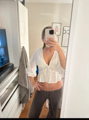 Vit blus med broderade blommor - Superfin vit blus med broderade blommor och v-ringning. Blusen har trekvartsärmar, knappar framtill och en lätt croppad. VIKTIGT: se sista bild för att se att knappen är lätt sönder men går enkelt att laga.