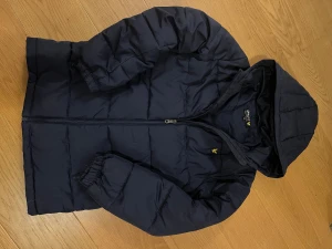 Mörkblå pufferjacka från Lyle & Scott - Lyle & Scott jacka – XS (13–14 år) Mörkblå pufferjacka från Lyle & Scott. Storlek 13–14 år men sitter som en XS. Jackan är i fint skick med ett litet hål på vänstra armen (syns knappt).  Varm, lätt och bekväm. Huvan och dragkedjan fungerar bra.  Perfekt till höst och vinter. 