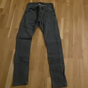 Grå Lee Luke jeans W27 L32 - Snygga grå jeans från Lee, modell Luke. De har en rak och smal passform med klassisk femficksdesign och dragkedja. Materialet är mjukt och slitstarkt denim i bomull. Perfekt för dig som gillar stilrena och moderna jeans.