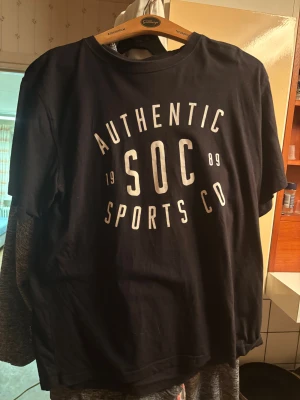 Svart t-shirt från SOC med tryck - Svart t-shirt från SOC i storlek XL med stort vitt tryck på bröstet där det står 'AUTHENTIC SOC SPORTS CO 1989'. Klassisk rund hals och korta ärmar. Skön och mjuk bomullskvalitet, perfekt för en sportig och avslappnad stil.