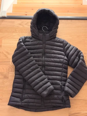 Svart pufferjacka från Everest - Svart pufferjacka från Everest med huva och dragkedja framtill. Jackan har quiltad design, två fickor med dragkedja och är lätt vadderad för extra värme. Perfekt för kyliga dagar och enkel att matcha med olika outfits.