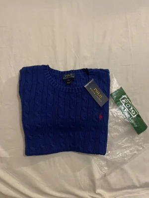 Blå kabelstickad tröja Polo Ralph Lauren - Blå kabelstickad tröja från Polo Ralph Lauren med rund hals och ribbade muddar. Tröjan har ett litet rött Polo-logo broderat på bröstet och är tillverkad i mjuk bomull. Perfekt för dig som gillar klassisk och stilren design.