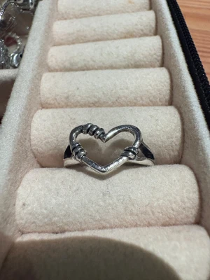 Hjärt ring - Unik silvrig ring med öppet hjärta i mitten, dekorerad med detaljer som ser ut som små lindningar runt hjärtat. Ringen är justerbar och har en cool, modern vibe som passar perfekt för dig som gillar statement-smycken.