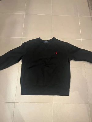 Svart sweatshirt från Polo Ralph Lauren - Svart sweatshirt från Polo Ralph Lauren i storlek M. Tröjan har ett klassiskt snitt med rund hals och långa ärmar. På bröstet finns den ikoniska röda Polo-loggan broderad. Materialet är mjuk bomull som känns skön mot huden. OBS sömmen har låstnat lite på vänster sida av bröstet. Men det syns inte så mycket