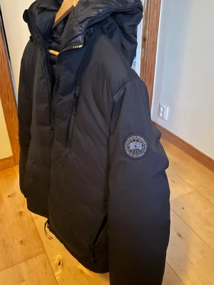 Svart dunjacka från Canada Goose - Säljer en svart dunjacka från Canada Goose med huva och diskret logga på ärmen. Jackan har dragkedja framtill, flera fickor och en klassisk pufferstil som håller dig varm under kalla dagar. Perfekt för dig som vill ha en stilren och funktionell vinterjacka.