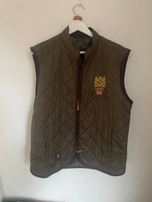 Olivgrön väst från Morris - Snygg quiltad olivgrön väst från Morris med guldbroderad logga och brittisk flagga på bröstet. Västen har dragkedja framtill, två fickor och är tillverkad i polyester. Perfekt för dig som gillar klassisk stil med moderna detaljer.