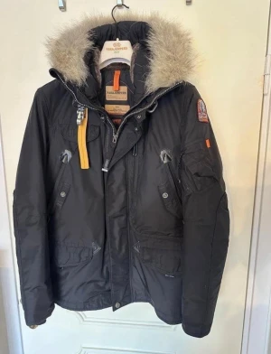Parajumpers svart vinterjacka med päls - Svart Parajumpers Right hand vinterjacka parkas med snygg päls på huvan, ikonisk gul rem och patch på ärmen. Jackan har flera fickor, dragkedja och knappar, samt quiltat foder. Endast använd en säsong och i nyskick!