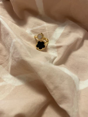 Guldfärgad ring med blommor - Unik guldfärgad ring med två blommor framtill, en svart och en vit. Ringen har en öppen design och blommorna har en mjuk, rundad form. Perfekt för dig som vill ha ett smycke som sticker ut och ger en cool touch till din stil.