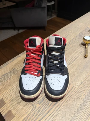 Nike Air Jordan 1 High röd/svart/vit - Nike Air Jordan 1 High sneakers i rött, svart och vitt med klassisk röd swoosh och svart/röd snörning. Skorna har rund tå, platt sula och högt skaft. Ovandelen är i skinn och mesh, med ikonisk Air Jordan-logga på sidan. Perfekt för dig som vill sticka ut med en retro basketstil.