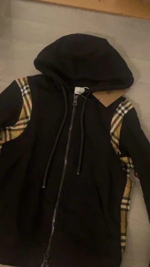 Burberry kofta - Svart hoodiejacka från Burberry med klassiska rutiga paneler i beige, rött och svart längs ärmarna. Jackan har huva med snörning och hel dragkedja framtill. Perfekt för dig som vill ha en streetig och lyxig look. 10/10