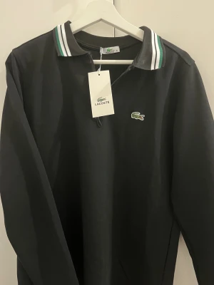 Svart pikétröja Lacoste med krage - Svart långärmad pikétröja från Lacoste med klassisk krage i svart, vit och grön. Tröjan har dragkedja vid halsen och den ikoniska krokodilloggan broderad på bröstet. Perfekt för dig som gillar stilrena och sportiga plagg.