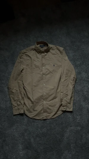 Ralph Lauren Skjorta Beige - En beige Ralph lauren skjorta som bara har provats, aldrig används! Bara legat och samlat damm i garderoben i 1,5 år, inga defekter av huvud taget förutom att tyget har förlorat lite färg på ett litet ställe men märks inte vid användning (syns på sista bilden).