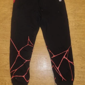 Svarta mjukisbyxor med rött mönster - Svarta mjukisbyxor med coolt rött grafiskt mönster på benen. Byxorna har muddar vid bensluten och är perfekta för chill eller träning. Materialet är mjukt och bekvämt, och designen sticker ut med sin unika stil.
