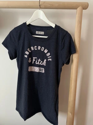 Mörkblå Abercrombie & Fitch t-shirt - Snygg mörkblå t-shirt från Abercrombie & Fitch med klassisk rund hals och tryckt logga i vitt och ljusrosa på bröstet. Modellen är kortärmad och har en normal passform. Materialet är mjuk bomull.