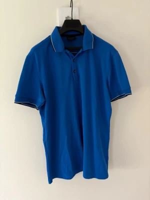 Blå golfpiké med vita detaljer från Galvin Green - Snygg blå pikétröja från Galvin Green med vita ränder på krage och ärmslut. Klassisk passform, kortärmad och med knappar framtill. Perfekt för en sportig look eller till golfbanan. Storlek: L passar M. Nypris: 999kr 