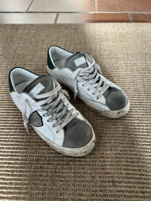 Philippe Model vita sneakers strl 37 junior  - Snygga vita sneakers från Philippe Model med grå mockadetaljer på tån och plös, grönt parti med gul kant bak på hälen och svarta sulor. Skorna har snörning och är tillverkade i skinn och mocka. ”Smuts” på sulorna är design.