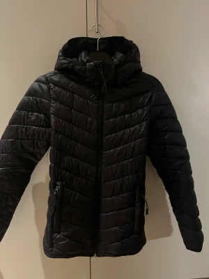 Svart tunnare jacka 146/152 - Säljer en svart jacka med huva och dragkedja framtill. Jackan har quiltad design med sneda sömmar och två fickor med dragkedja. 