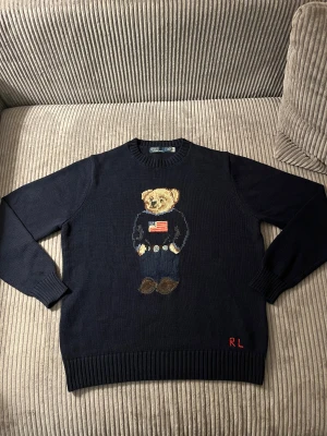 Mörkblå stickad tröja Polo Bear - Säljer en mörkblå stickad tröja från Polo Ralph Lauren med ikonisk Polo Bear broderad på bröstet och RL-broderi i rött vid nederkanten. Tröjan har rund hals, ribbade muddar och är långärmad. Perfekt för dig som gillar klassisk stil med en twist.