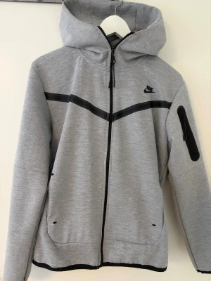 Grå Nike vindjacka med huva - Snygg grå vindjacka från Nike med svart logga på bröstet och svarta detaljer längs bröstet. Jackan har hel dragkedja, huva och praktisk ficka med dragkedja på ärmen. Perfekt för dig som gillar sportig och clean stil. Är liten i storleken. 