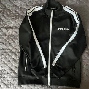 Svart track jacket från Palm Angels - Kommer perfekt passa till sommaren. Säljer en svart track jacket från Palm Angels med vita ränder längs ärmarna och vit dragkedja. Perfekt för en sportig och trendig look.