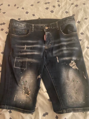 Mörkblå jeansshorts Dsquared2 ICON - Säljer ett par mörkblå jeansshorts från Dsquared2 ICON med slitna detaljer, färgstänk och coola tvättade effekter. Klassisk femficksmodell med knapp och dragkedja framtill. Märkespatch baktill och råa kanter för extra edge.
