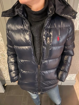 Mörkblå dunjacka från Ralph Lauren - Snygg mörkblå dunjacka från Ralph Lauren med glansig finish, huva och dragkedjor. Jackan har röd broderad logga på bröstet och en bröstficka med RL-dragkedja. Perfekt för kalla dagar och riktigt stilren look.