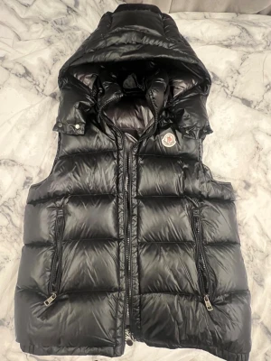 Svart dunväst från Moncler med huva - Svart dunväst från Moncler med glansig finish, stor vadderad huva och två fickor med dragkedja. Klassisk Moncler-logga på bröstet och quiltad design. Perfekt för lager-på-lager och streetstyle. Stängs med dragkedja framtill.