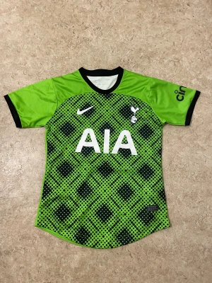 Grön Tottenham Hotspur fotbollströja - Säljer en grön Tottenham Hotspur fotbollströja från Nike med svart och vit detaljer. Tröjan har korta ärmar, svart krage och är tillverkad i lätt polyester. Framsidan har ett grafiskt mönster med svarta prickar och rutor, samt klubbmärke och AIA-logga.
