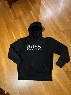 Hugo Boss Hoodie - Svart hoodie från Hugo Boss med stor vit logga på bröstet. Perfekt för dig som gillar stilrena och bekväma plagg. Hör av dig vid minsta fundering!:)