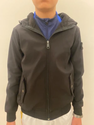 Svart vindjacka från Stone Island - Svart vindjacka från Stone Island med huva och ikonisk patch på vänster ärm. Jackan har dragkedja framtill, två fickor med knappstängning och ribbade muddar. Materialet är syntet och jackan har en loose passform som är chill och enkel att bära.