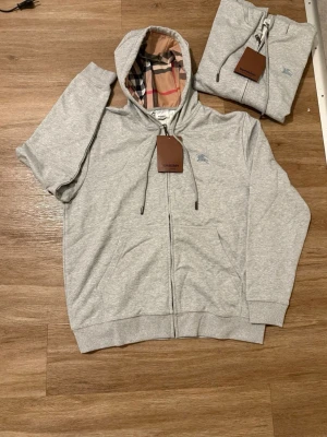 Grå hoodie med rutigt foder, stl M – Nytt skick - Snygg och stilren grå hoodie i storlek M, i helt nytt skick. Tröjan har dragkedja framtill och ett mjukt, mönstrat innerfoder som gör den både bekväm och unik i stilen. Passar perfekt till vardags, både ensam eller under en jacka.