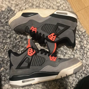 Nike Air Jordan 4 Retro Grå/Orange - Nike Air Jordan 4 Retro sneakers i grå mocka med svarta och orange detaljer. Skorna har klassisk snörning, meshpaneler på sidorna och en vit platt sula med synlig Air-enhet. Ikonisk Jumpman-logga på plösen och orange insida för extra pop.