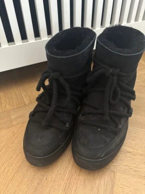 Svarta moon boots från Inuikii - Moon boots från Inuikii i helsvart mocka med grova snören och fluffigt foder. Skorna har rund tå, platt sula och högt skaft som håller dig varm. Perfekt för kalla vinterdagar och har en cool, chunky look som sticker ut.