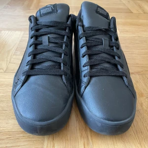 Svarta sneakers från Puma - Snygga svarta sneakers från Puma med klassisk design och rund tå. Skorna har snörning och är tillverkade i skinn med detaljer i syntet. Puma-loggan syns både på plösen och hälen. Perfekta för dig som gillar stilrena och sportiga skor.