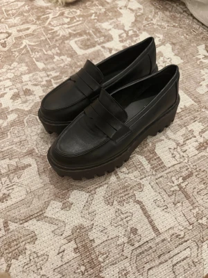 Chunky svarta loafers med plattform - Säljer ett par svarta loafers med riktigt chunky plattformssula och grovt mönstrad undersida. Skorna har klassisk pennyloafers-design med rund tå och är tillverkade i skinn. Perfekta för dig som gillar att sticka ut med din stil.