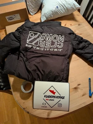  Moncler jacka  - En Svart Special pufferjacka från Moncler med stora vita tryck på ryggen och bröstet, samt logga på ärmen. Jackan har två dragkedjeförsedda fickor fram och ribbade muddar vid ärmslut. Perfekt för dig som gillar streetwear och vill sticka ut med en fet design. Vattentät och vattentät mobilficka