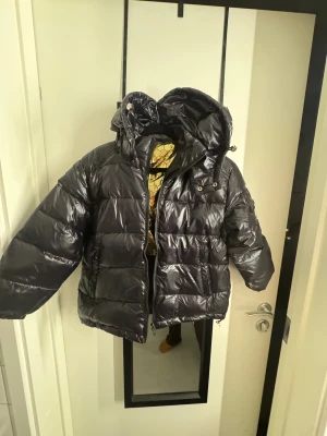 Svart Moncler pufferjacka med huva - Säljer en svart pufferjacka från Moncler med glansig finish, huva och dragkedja. Jackan har Moncler-logga på ärmen, praktiska fickor och coolt guldigt innerfoder med hexagonmönster. Insidan har även en serietidningsdetalj. Perfekt för kalla dagar. Stl 130 tycker jag men är märkt 140. Därför säljer vi för det blev fel. 
