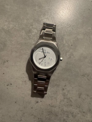 ”Who cares I’m already late” klocka - Säljer en silvrig klocka med metallarmband och vit urtavla. Urtavlan har handskrivna siffror och texten 'Who cares I’m already late' vilket ger en skön och avslappnad vibe. Klockan väger 132 gram och har ett klassiskt lås på armbandet.