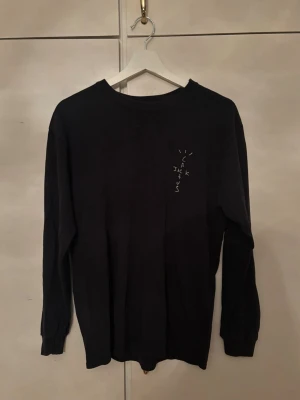 Svart långärmad Cactus Jack Jackboys topp - Svart långärmad topp från Cactus Jack med tryck på bröstet och ett stort Jackboys-motiv med sprucken glasdesign på ryggen. Rund hals och mjukt bomullsmaterial som känns skönt mot huden. Perfekt för dig som gillar streetwear och Travis Scott.