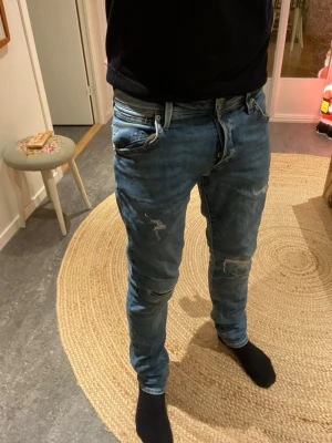 Blå slitna jeans från Jack & Jones - Säljer ett par blå jeans från Jack & Jones med snygga slitningar och lagom tvättad look. Modellen är smalare i benen och har klassiska fem fickor samt dragkedja. Perfekta för dig som gillar en avslappnad men trendig stil.